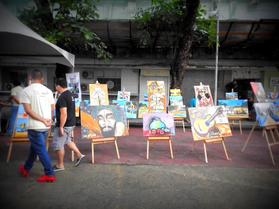 Exposição de arte ao ar livre com várias pinturas em cavaletes e pessoas caminhando pelo local.