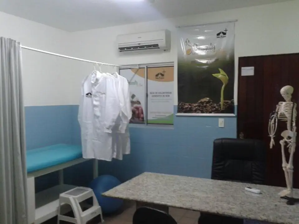 Sala de atendimento com maca, jalecos pendurados e modelo anatômico de esqueleto ao lado da mesa.