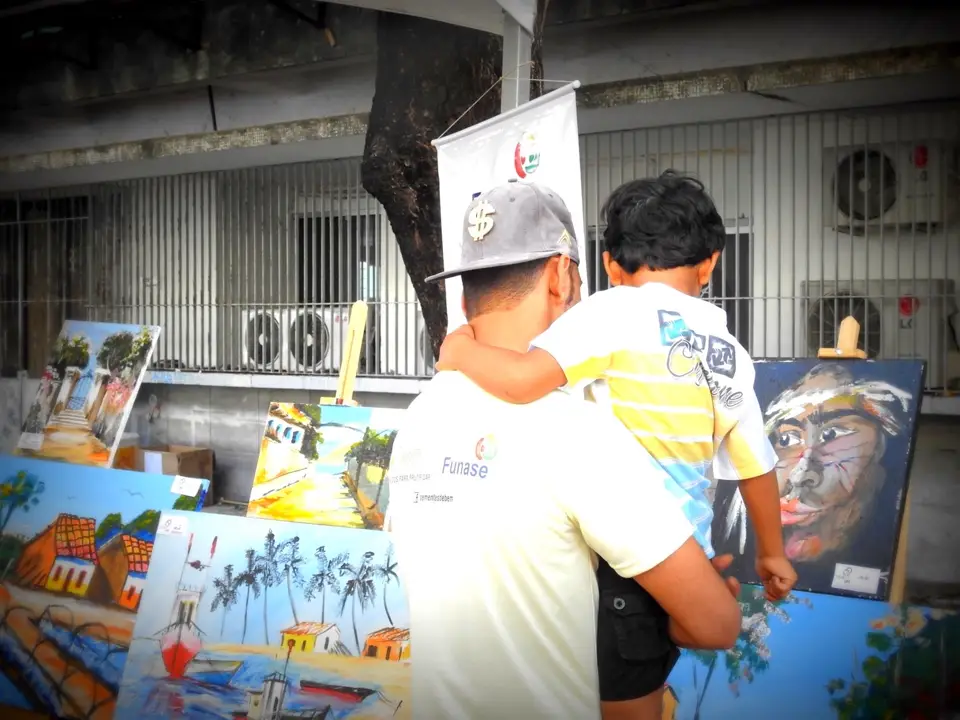 "Homem com uma criança no colo observa pinturas em cavaletes numa exposição ao ar livre.