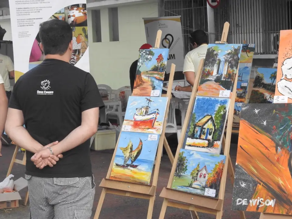 Visitante observa quadros em cavaletes durante uma exposição de arte comunitária do projeto 99 Por Um